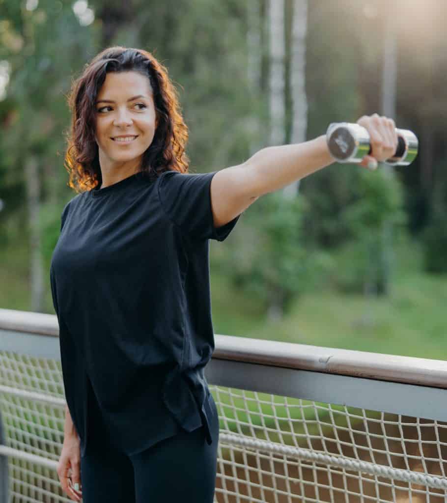 Active energized sportswoman stretches arms with dumbbell, poses on bridge over nature background