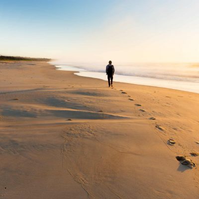 A person walking on a beautiful beach leaves a trail of footprints as the sunrise illuminates mist rising from the sea in golden light.